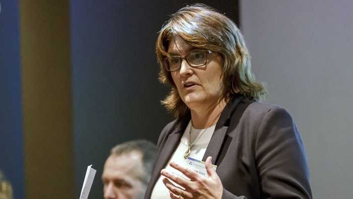 A Caucasian woman with brown hair, wearing glasses and a blazer.