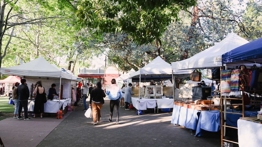 Market stalls under large green trees and two people walking in the middle of the image