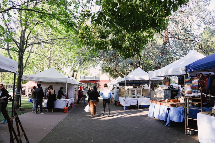 Market stalls under large green trees and two people walking in the middle of the image