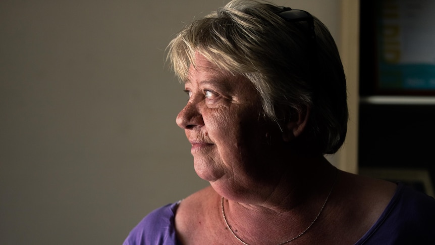 A woman with short blonde hair looks out of a window of a dimly lit room.