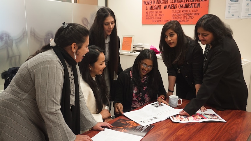 Six women gather around a table, looking at magazines.