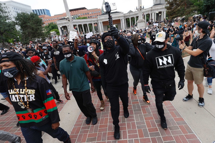 A man holds his mobile device to record a march as crowds of people walk behind him.