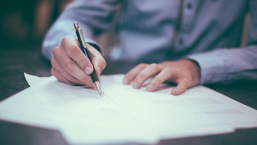 A photo of a man's hands signing a document. He wears a blue business shirt. 