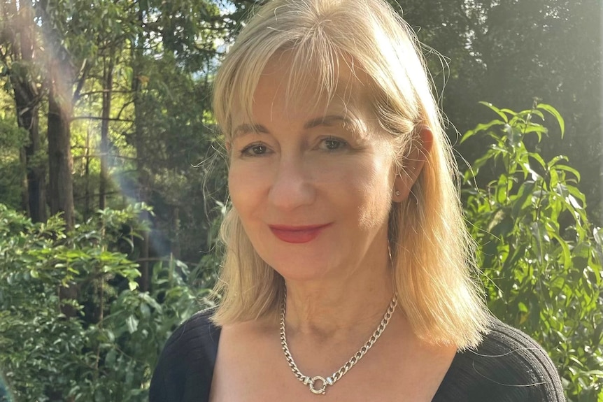 A blonde woman wearing a black shirt, stands on a balcony near bushland, smiling.