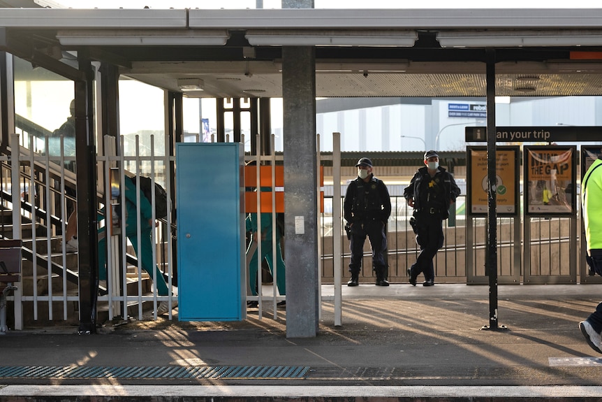 Deux policiers se tiennent à l'arrière d'un quai de train.  Plusieurs adolescents en uniforme scolaire passent.