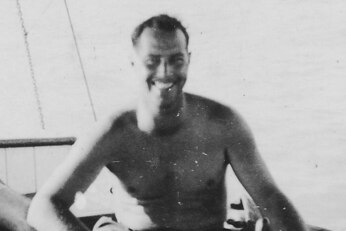 A man not wearing a shirt smiles at the camera in a black and white photo.