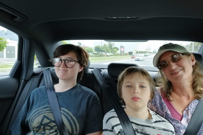 A mother wearing glasses and a hat sits with her teenage daughter and son in the back of a car