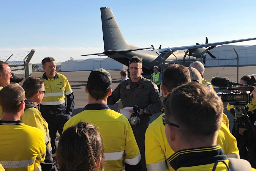 A group of people in yellow trades shirts is briefed by an air force officer in front of a large RAAF plane.