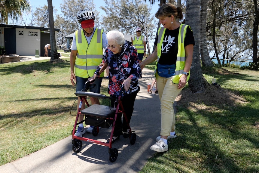 Mary and another volunteer wearing high vis, walking beside Thelma, 104, who is using her walking frame.