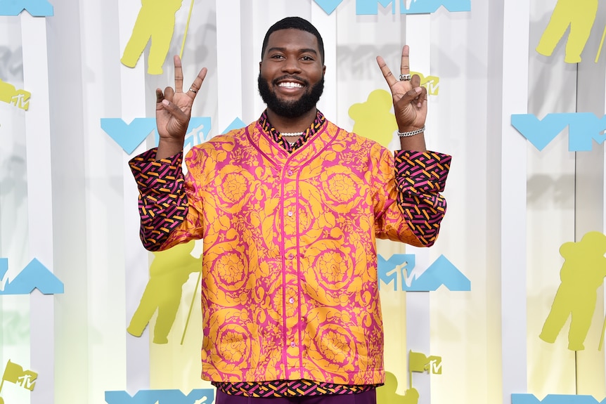khalid smiles and poses holding both his hands in peace signs on an arrivals carpet wearing an orange and pink shirt
