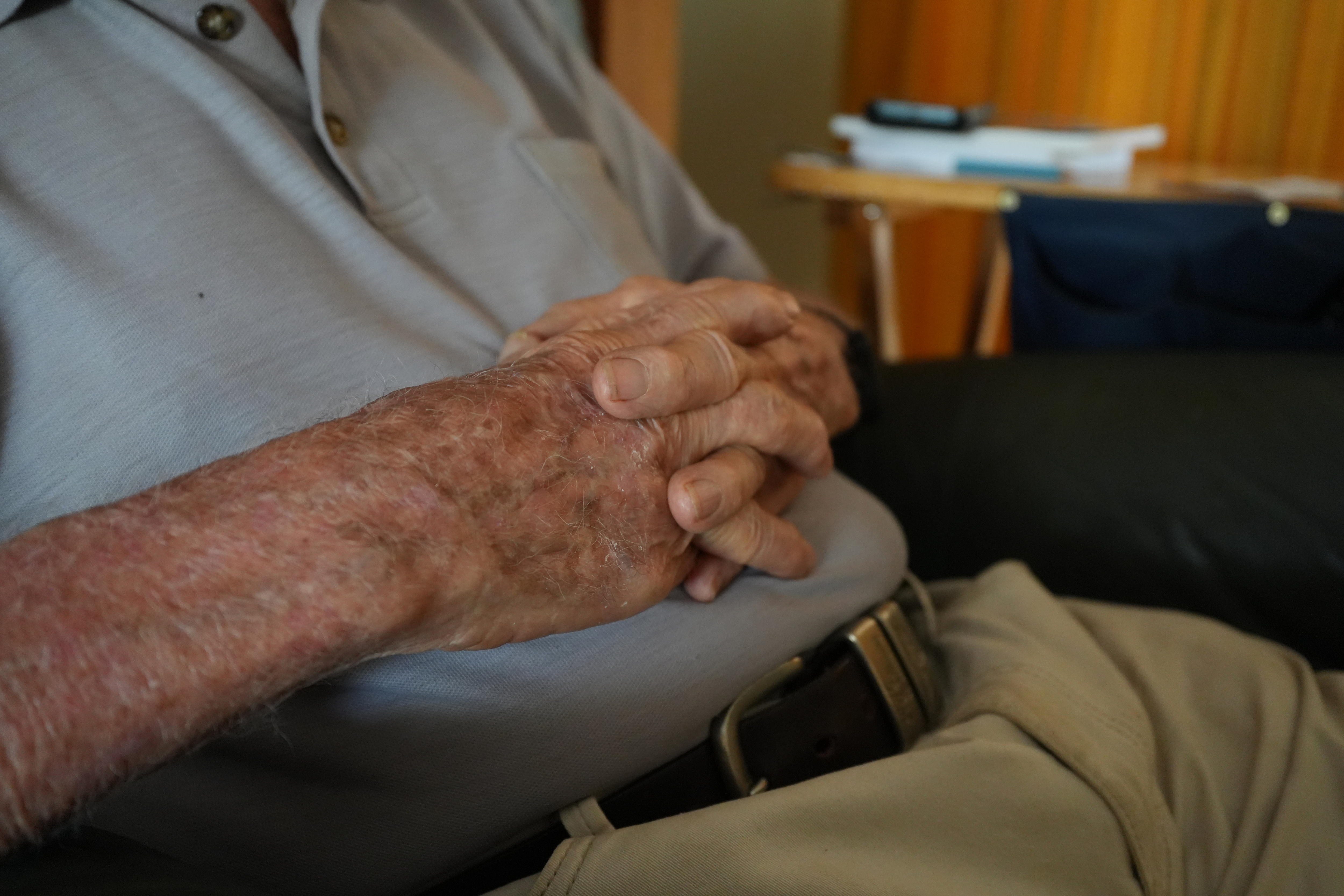 As mãos de Clarence 'Clack' John repousam sobre sua barriga. Ele está vestindo uma camisa pólo cinza, calça branca e faixa marrom.