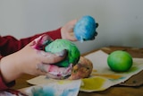 A little boy holds a green blob of plasticine in one hand and a blue blob in the other.