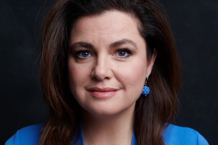 A white woman with long dark brown hair wearing a blue suit and matching blue earrings