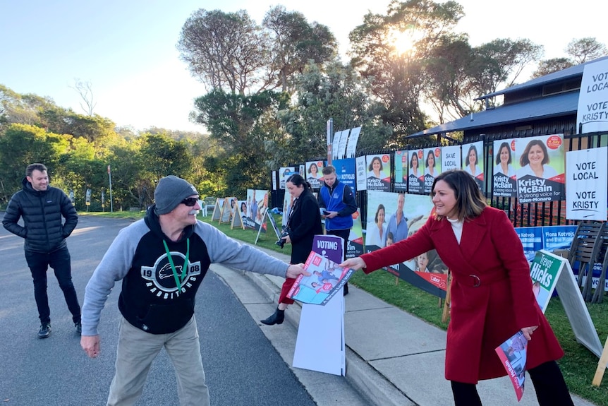 A woman in a red coat hands a piece of paper to a man in a beanie and sunglasses outside a polling station.