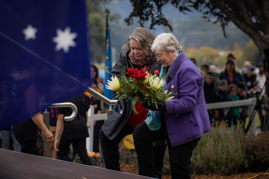 Two older women who look sad step up to a war memorial to lay flowers. 