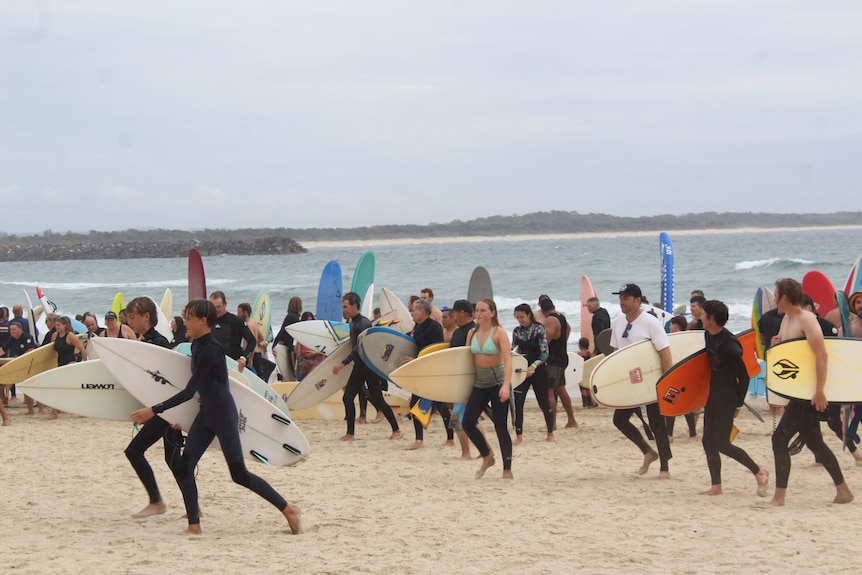 A large number of surfers, wearing wetsuits, carry their boards along a beach.