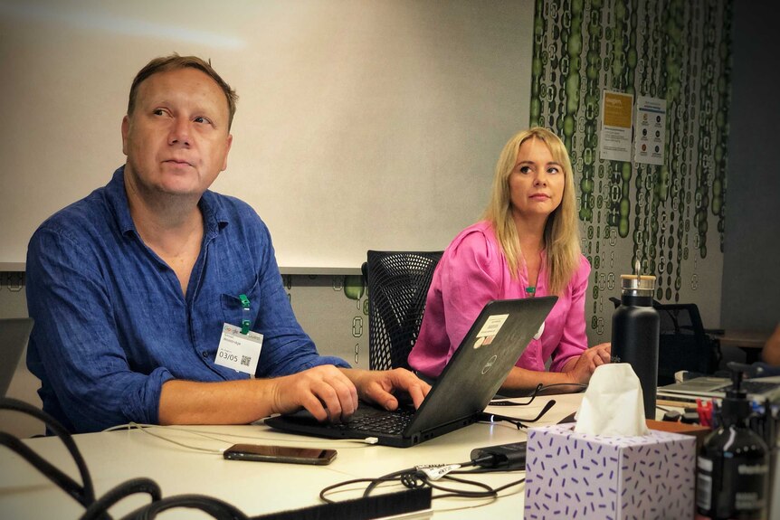 Two people sit at a desk in front of laptops; on the left a man and on the right, Anne Kruger.