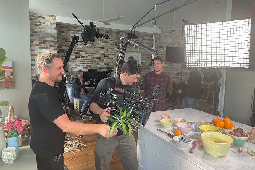 Camera crew filming food on a kitchen bench with a large light in the background.