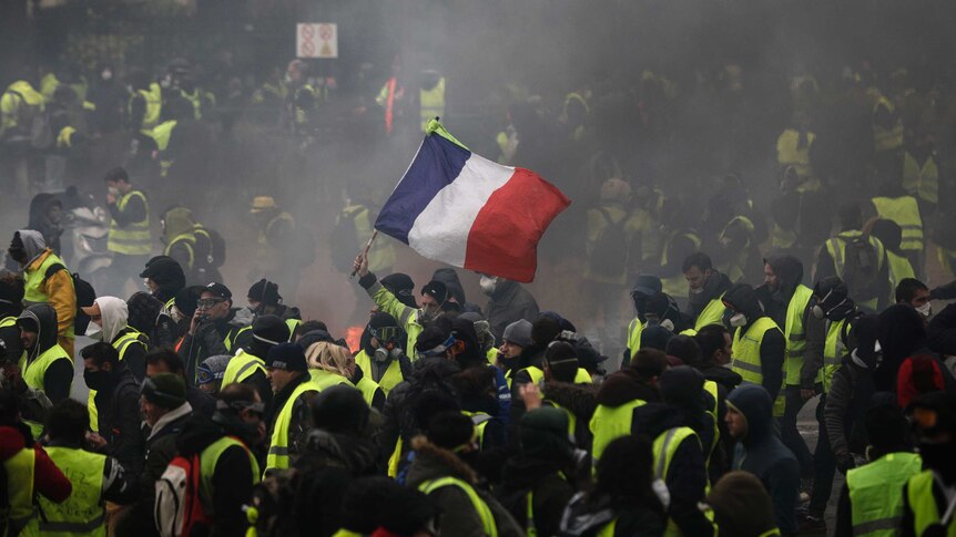 Hundreds of protesters wearing yellow vests gather in a Paris street, one holds up the French flag