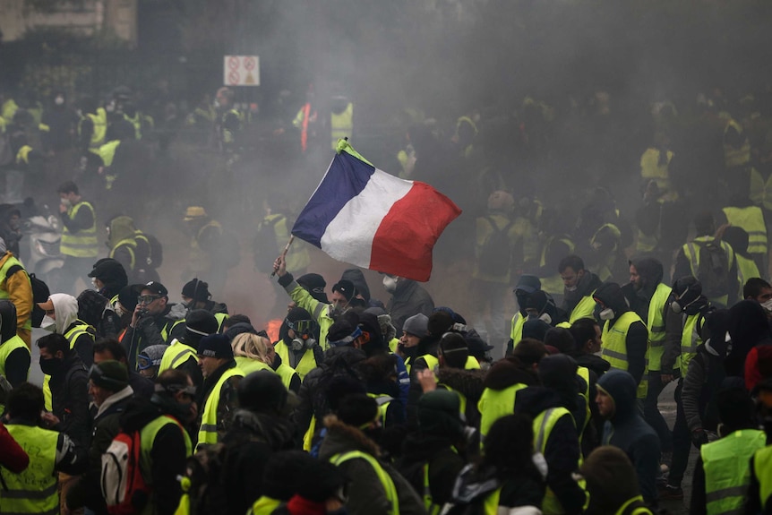 Hundreds of protesters wearing yellow vests gather in a Paris street, one holds up the French flag