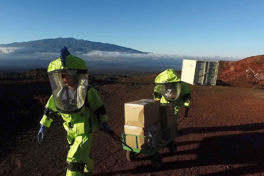 Two crew members in space suits pull a trolley up a volcano in Hawaii.