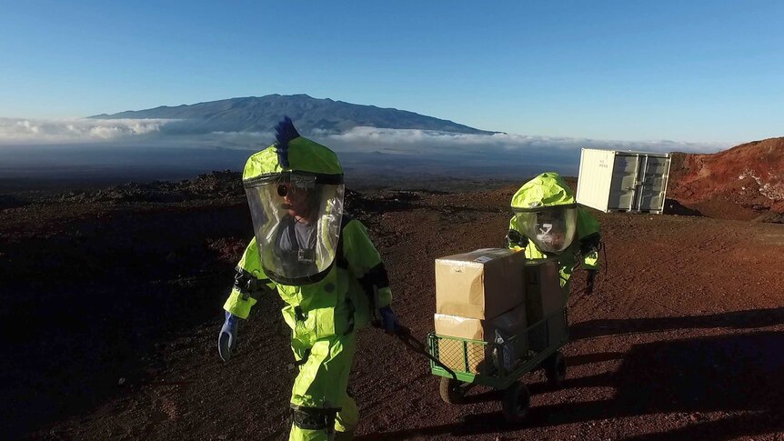 Two crew members in space suits pull a trolley up a volcano in Hawaii.