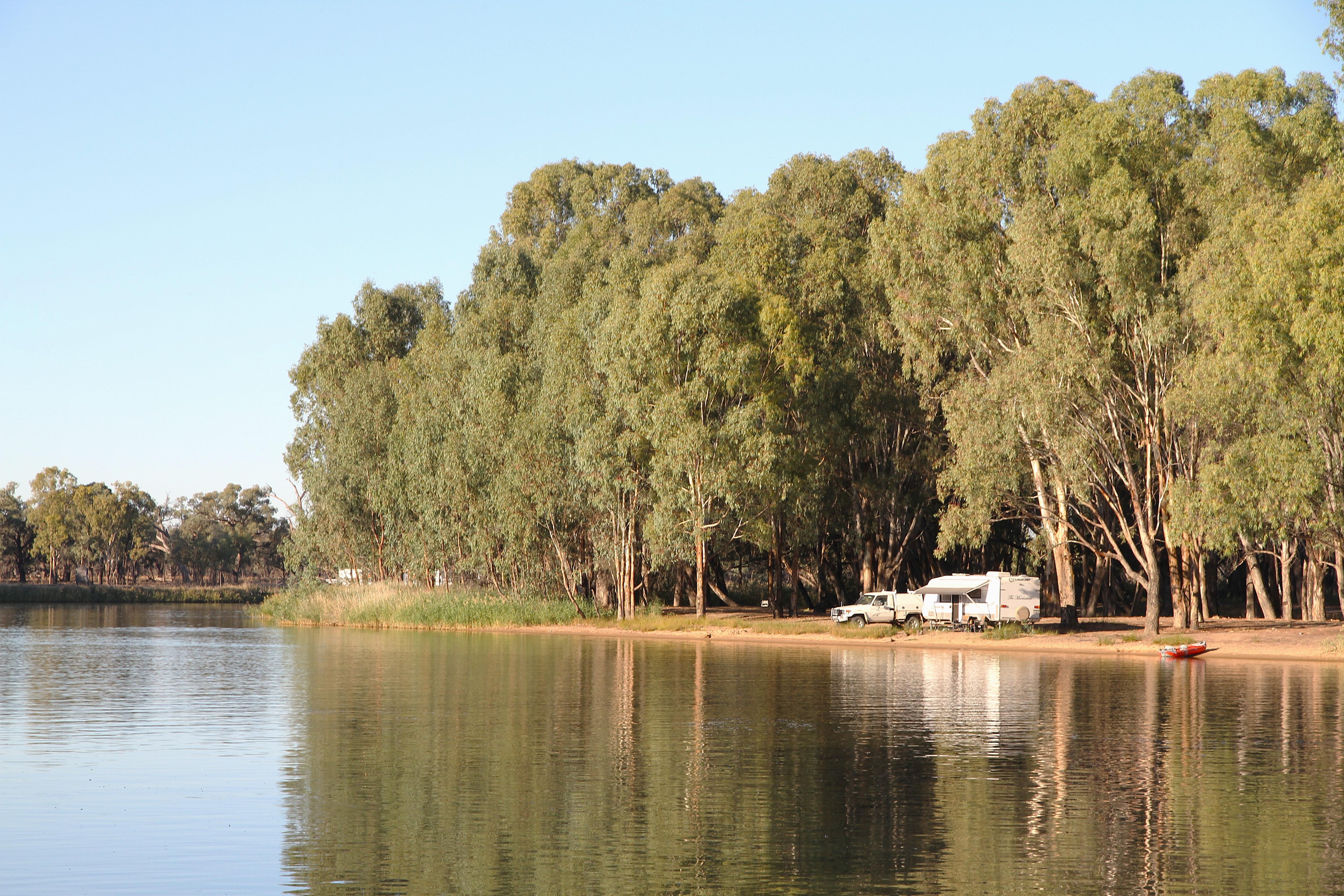 Un coche y una caravana están aparcados en un recodo del río, sobre un ancho río rodeado de altos y frondosos árboles verdes.