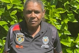 An Indigenous woman stands in front of a green hedge, straight-faced. 