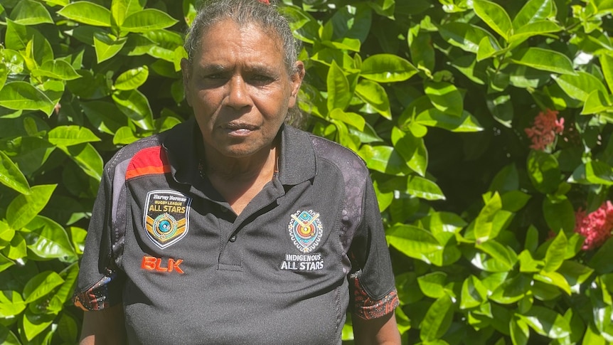 An Indigenous woman stands in front of a green hedge, straight-faced. 