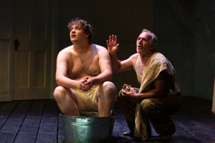 A young shirtless man in an old bathtub, an older man is speaking to him - all on stage