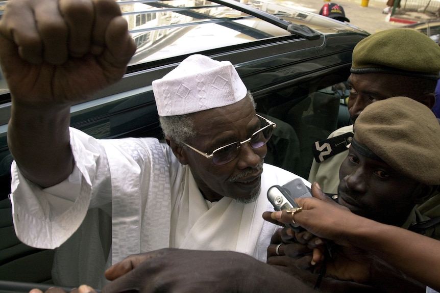 A close up of Hissene Habre getting out of a car surrounded by a media scrum. 