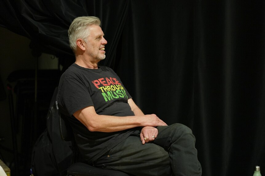 Stuart Davis is seated in front of a black curtain. He wears a black t-shirt that says 'peace through music'.