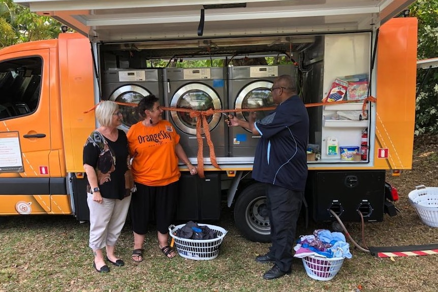 Two women and Aboriginal man cut ribbon in front of orange laundry van