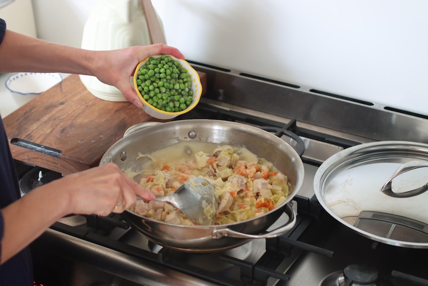 Peas being added to a saucepan of chicken and vegetables on a stovetop.