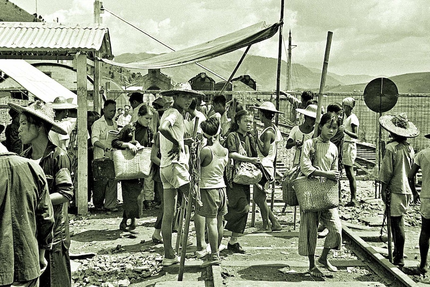 A black and white image shows a group of Chinese people wearing 1940s clothes in front of a border fence.