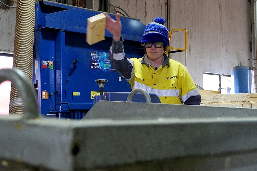 A person in a high-vis top and beanie throws a piece of wood inside a workshop. 
