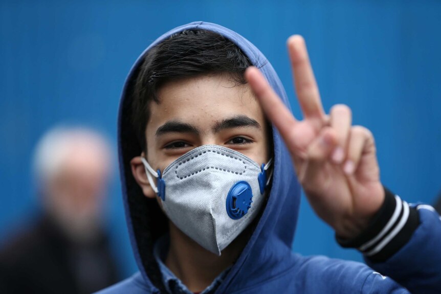 A young Iranian boy in a face mask gives a peace sign to the camera