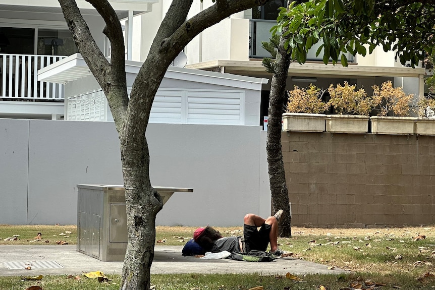 Homeless man lays in park with homes in background.
