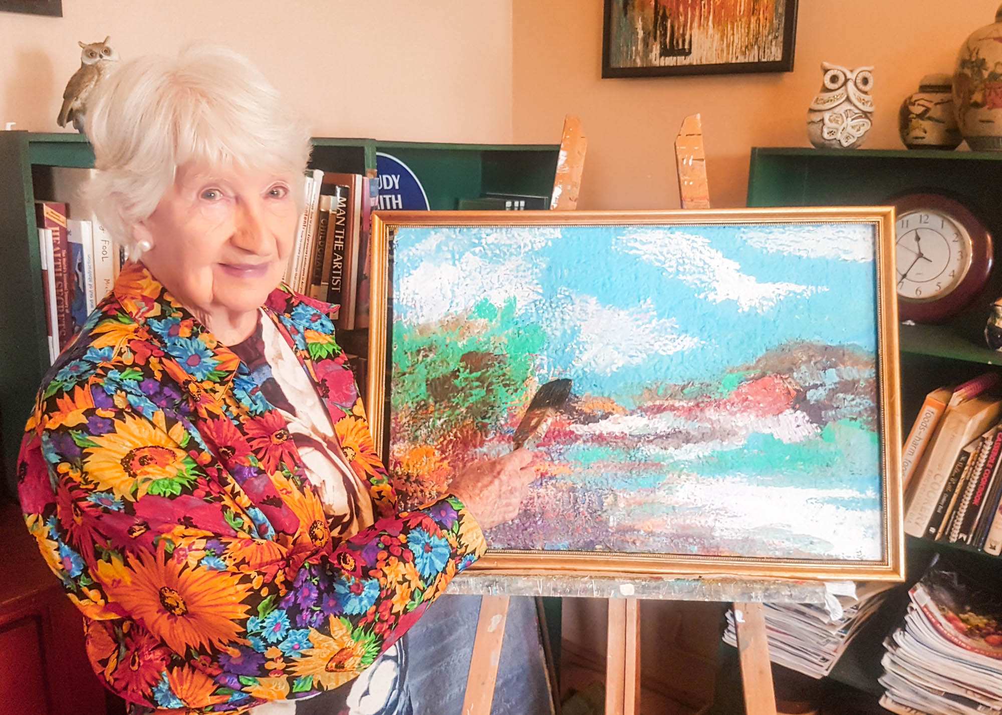 A senior woman with a flower shirt holding a paint brush standing in front of artwork on an easel in a golden frame.