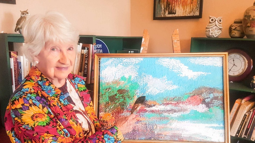 A senior woman with a flower shirt holding a paint brush standing in front of artwork on an easel in a golden frame.