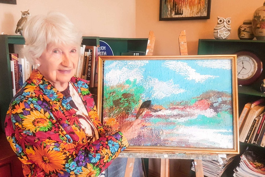 A senior woman with a flower shirt holding a paint brush standing in front of artwork on an easel in a golden frame.