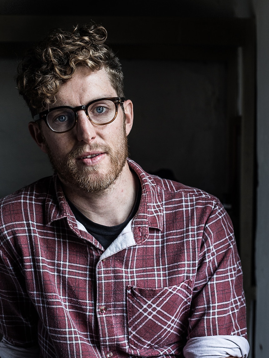 A bespectacled man with short strawberry blonde curly hair wears button up maroon plaid shirt and poses indoors near window.