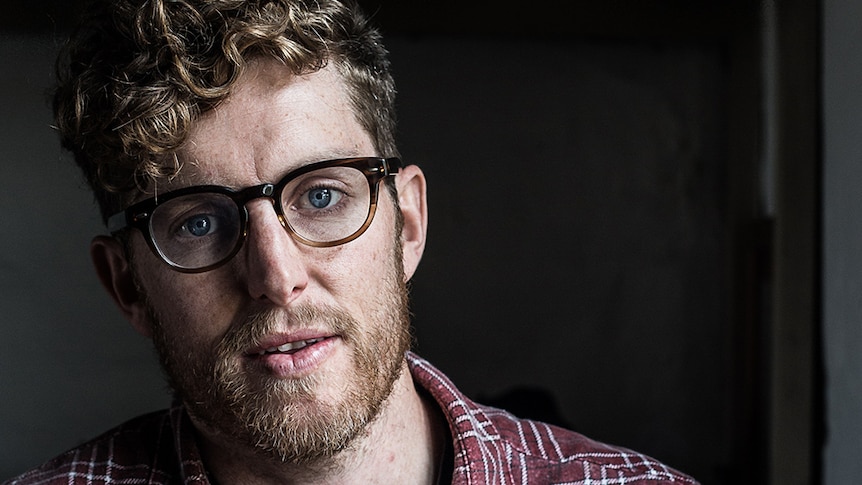 A bespectacled man with short strawberry blonde curly hair wears button up maroon plaid shirt and poses indoors near window.