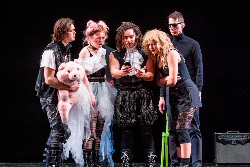 Five actors on stage; four of them in costume as members of a punk protest band, all looking at a phone