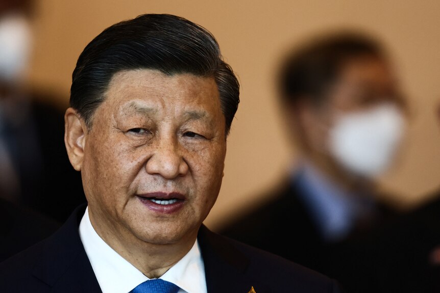 A close up of Xi Jinping smiling with a man wearing a mask standing behind him.