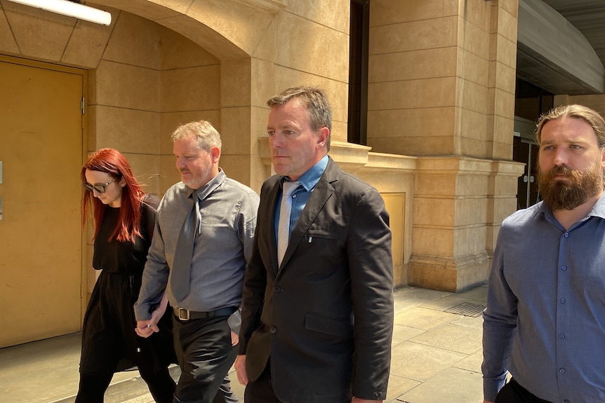 Three men in suits and a woman in black dress walk outside a court building