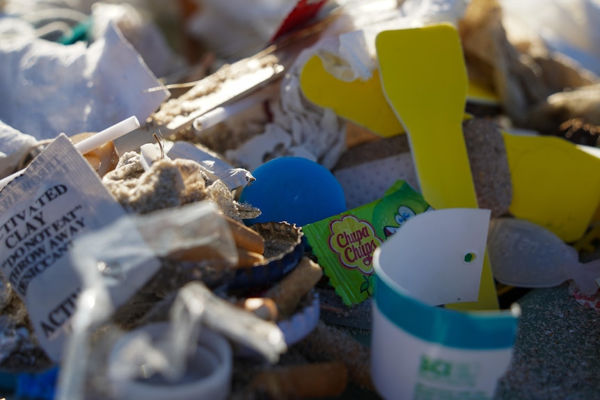 Chupa chup wrapper, ice cream cup and spoon, plastic and other rubbish in beach sand.