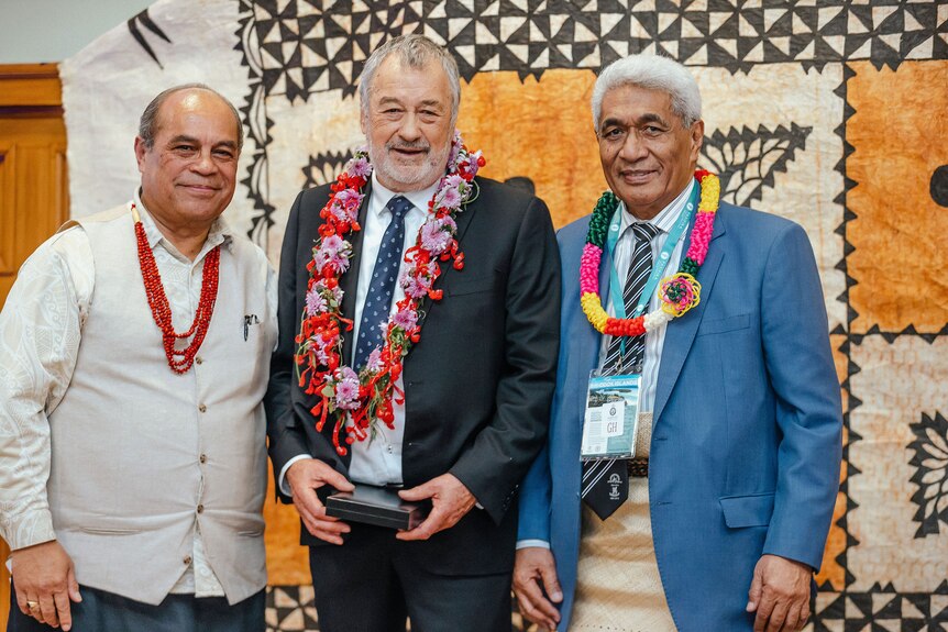 Three Pacific Island elder men wearing suits adorned by leis at a ceremony in front of orange, white and black tapa cloth 