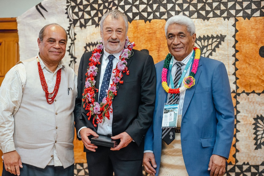 Three Pacific Island elder men wearing suits adorned by leis at a ceremony in front of orange, white and black tapa cloth 
