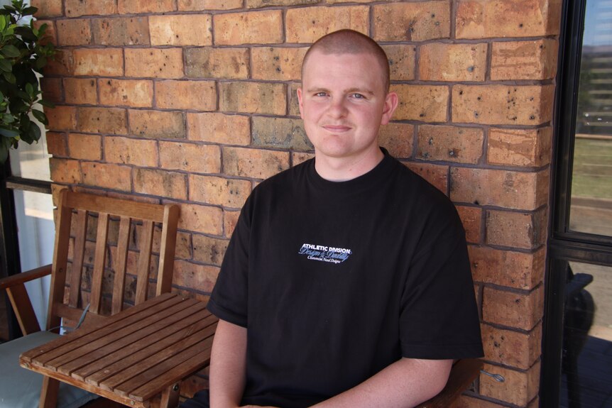 A young man with a shaved head sitting on a chair in front of a brick wall